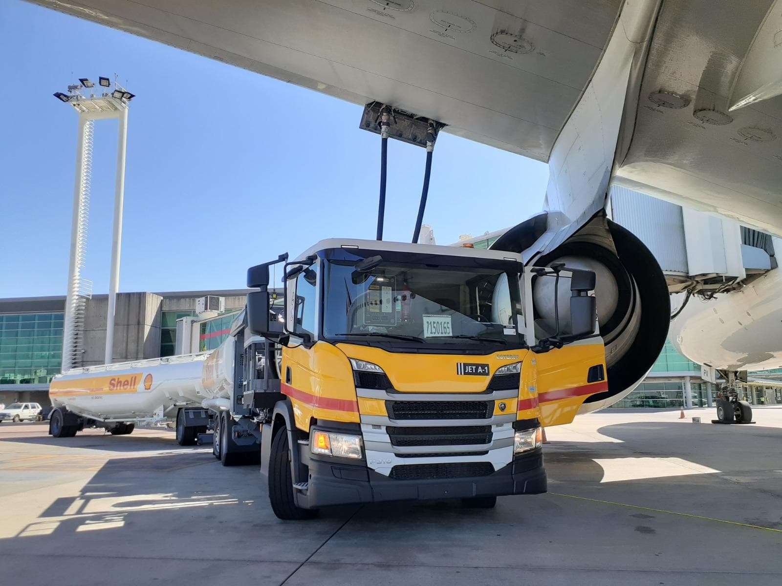 Un camión cisterna de Shell con combustible Jet A-1 carga un avión en la plataforma de un aeropuerto, con las mangueras conectadas debajo del ala.