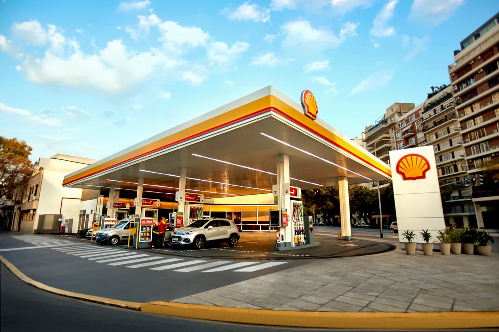 Una estación de servicio Shell de día, con autos cargando combustible bajo el techo y edificios de departamentos detrás.