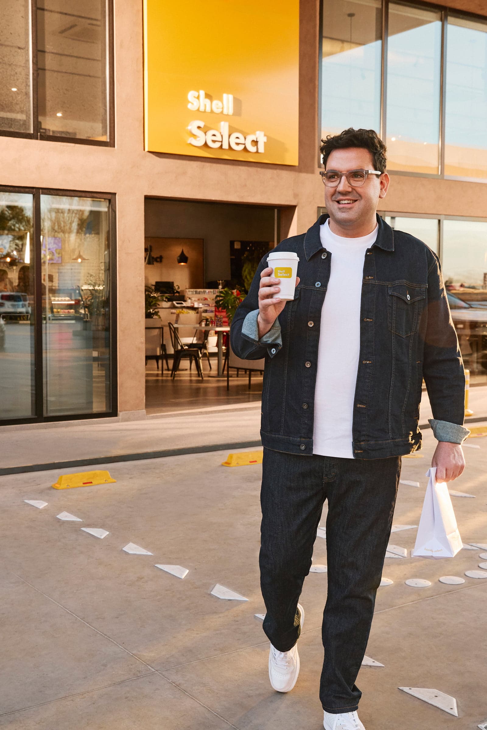 Un hombre sonriente sale de una tienda Shell Select con un café y una bolsa de compras.