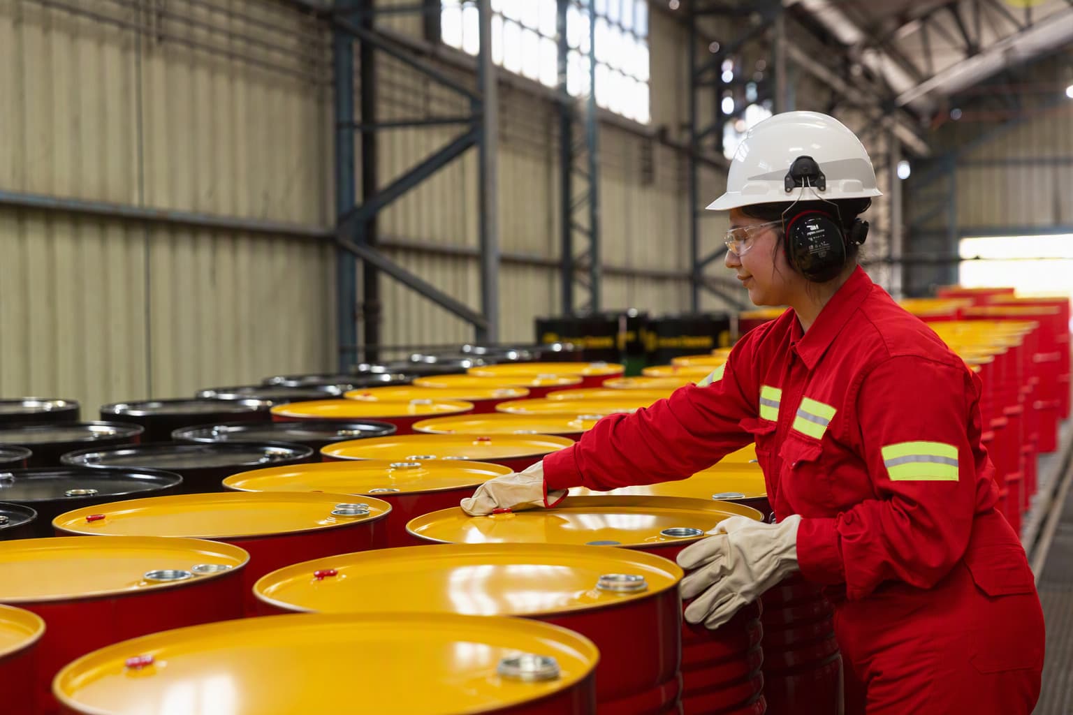 Una operaria con casco, protectores auditivos y mameluco rojo revisa tambores de lubricante Shell en la planta.